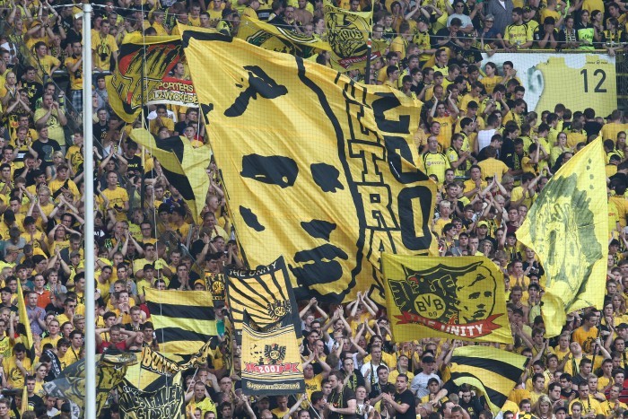 Man sieht frotal im Zoom den Block drölf der Südtribüne voll mit Fans und schwarz-gelben Fahnen. Die größte Fahne in der Mitte zeigt ein Konterfei von August Lenz und den Schritfzug "2001 Ultras"